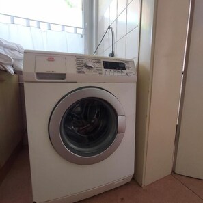 Lavadora de carga frontal con panel de control, ubicada en una esquina de una habitación con pared de azulejos y una ventana.