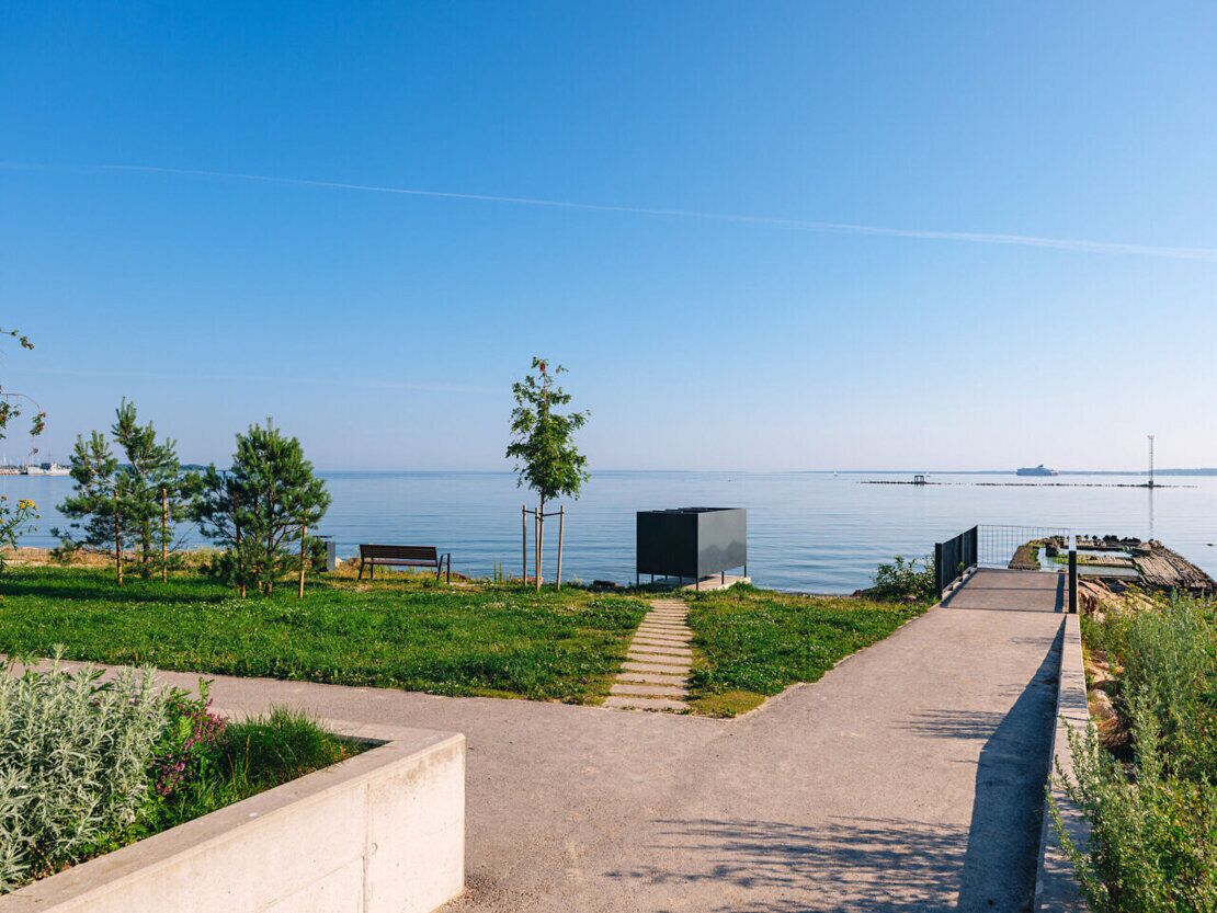A paved pathway leading to a body of water with a bench and a structure on the left.