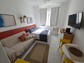 A compact hotel room with a bed, a sofa, a desk with a chair, a television, and a window with curtains. - Studio Pé na Areia em Copacabana (Rio de Janeiro)