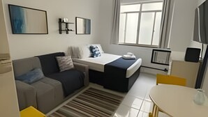 A modern bedroom with a bed, a sofa, a dining table, and a ceiling fan. - Studio Pé na Areia em Copacabana (Rio de Janeiro)