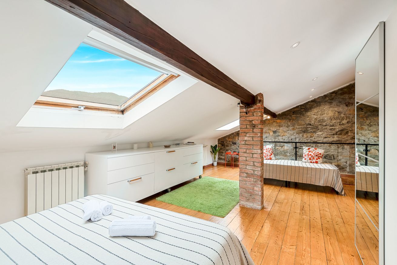 Ein Schlafzimmer mit Oberlicht, einem Bett mit gestreifter Bettdecke, einer weißen Kommode, einem grünen Teppich und einer Steinwand.