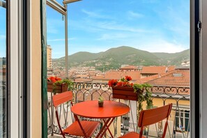 Ein Balkon mit Tisch und Stühlen, der auf einen Stadtblick mit Gebäuden und einer Hügelkulisse blickt.