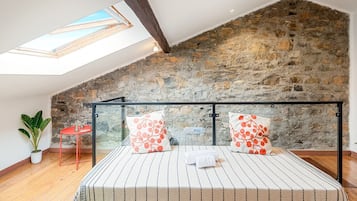 Une chambre avec un plafond en pente, un puits de lumière, un mur en pierre, un lit avec une literie à rayures et une chaise rouge.