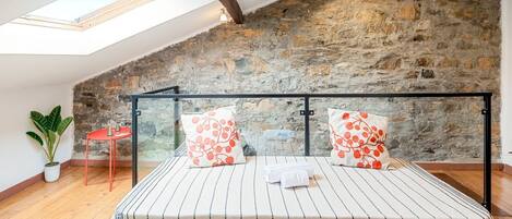 Une chambre avec un plafond en pente, un puits de lumière, un mur en pierre, un lit avec une literie à rayures et une chaise rouge.