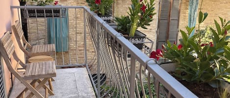 Un balcón con baranda de metal, sillas de madera, plantas en macetas y una pared de piedra.