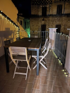 Une terrasse extérieure avec une table et des chaises en bois, une rambarde en métal et un bâtiment en briques en arrière-plan.