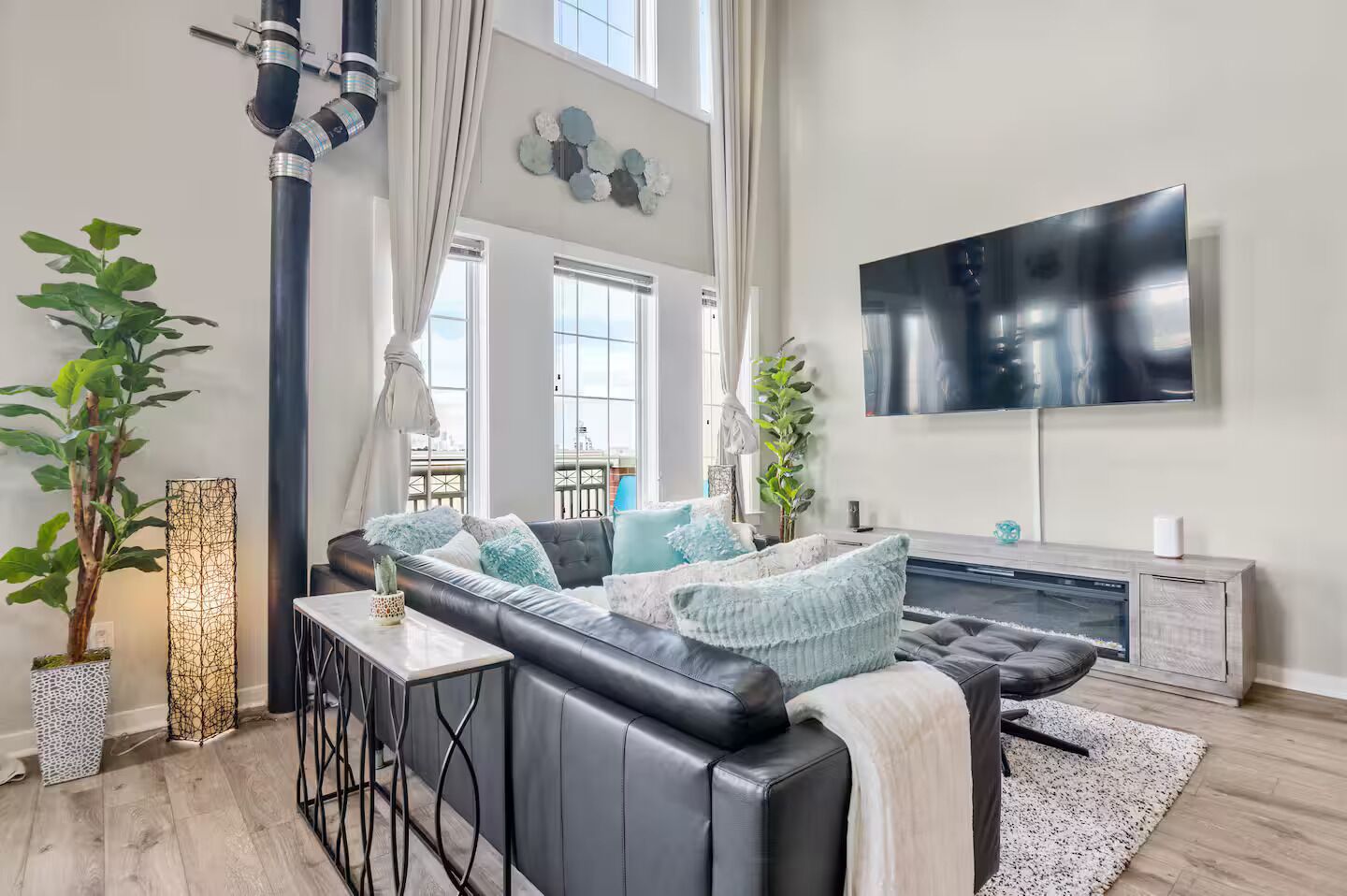 A modern living room with a black leather sofa, a flat-screen TV mounted on the wall, and a large window providing a view of the city.