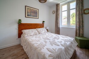 Ein Schlafzimmer mit hölzernem Kopfteil, einem Bett mit blumigem Überwurf, einem Gemälde an der Wand, einem Fenster mit Vorhängen und einer kleinen Pflanze auf dem Nachttisch.