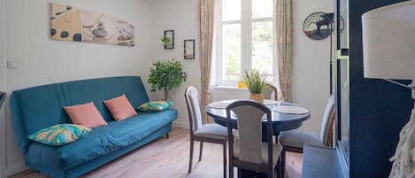 Uma sala de estar com um sofá azul, uma mesa de jantar com cadeiras, uma planta em vaso e uma obra de arte na parede.