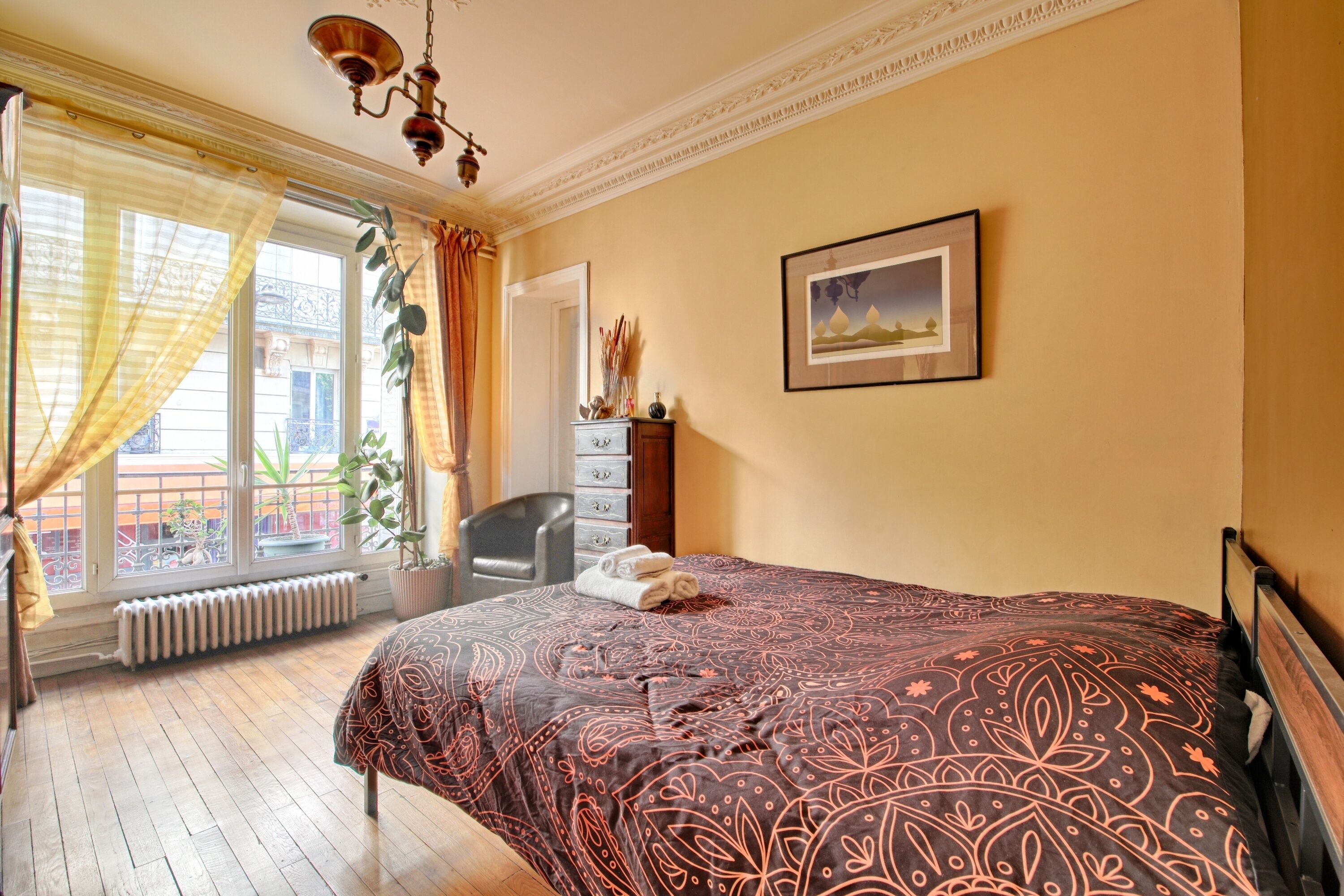A hotel room with a bed, a chair, a radiator, a window with curtains, and a painting on the wall.