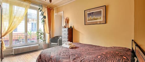 A hotel room with a bed, a chair, a radiator, a window with curtains, and a painting on the wall.