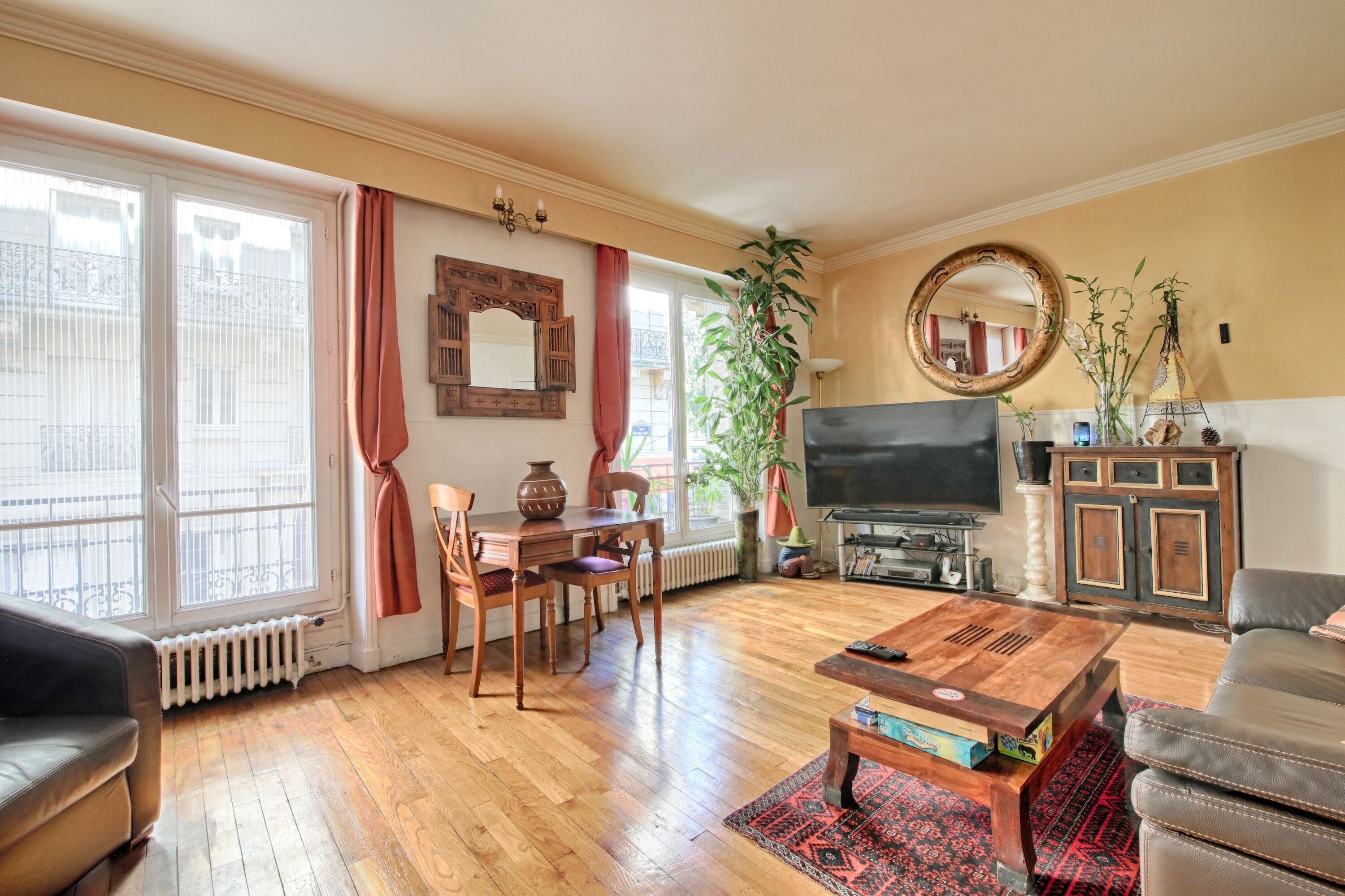 Una sala de estar con televisor, muebles de madera, un espejo grande y una ventana con cortinas.