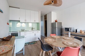 A modern kitchen with white cabinets, a stainless steel refrigerator, and a dining area with a round table and chairs. - Le Maryland - Climatisé à 100 m de la plage (Leucate)