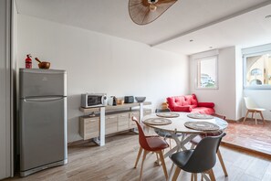 A compact kitchen with a refrigerator, microwave, and toaster, a dining area with a round table and chairs, and a red sofa. - Le Maryland - Climatisé à 100 m de la plage (Leucate)
