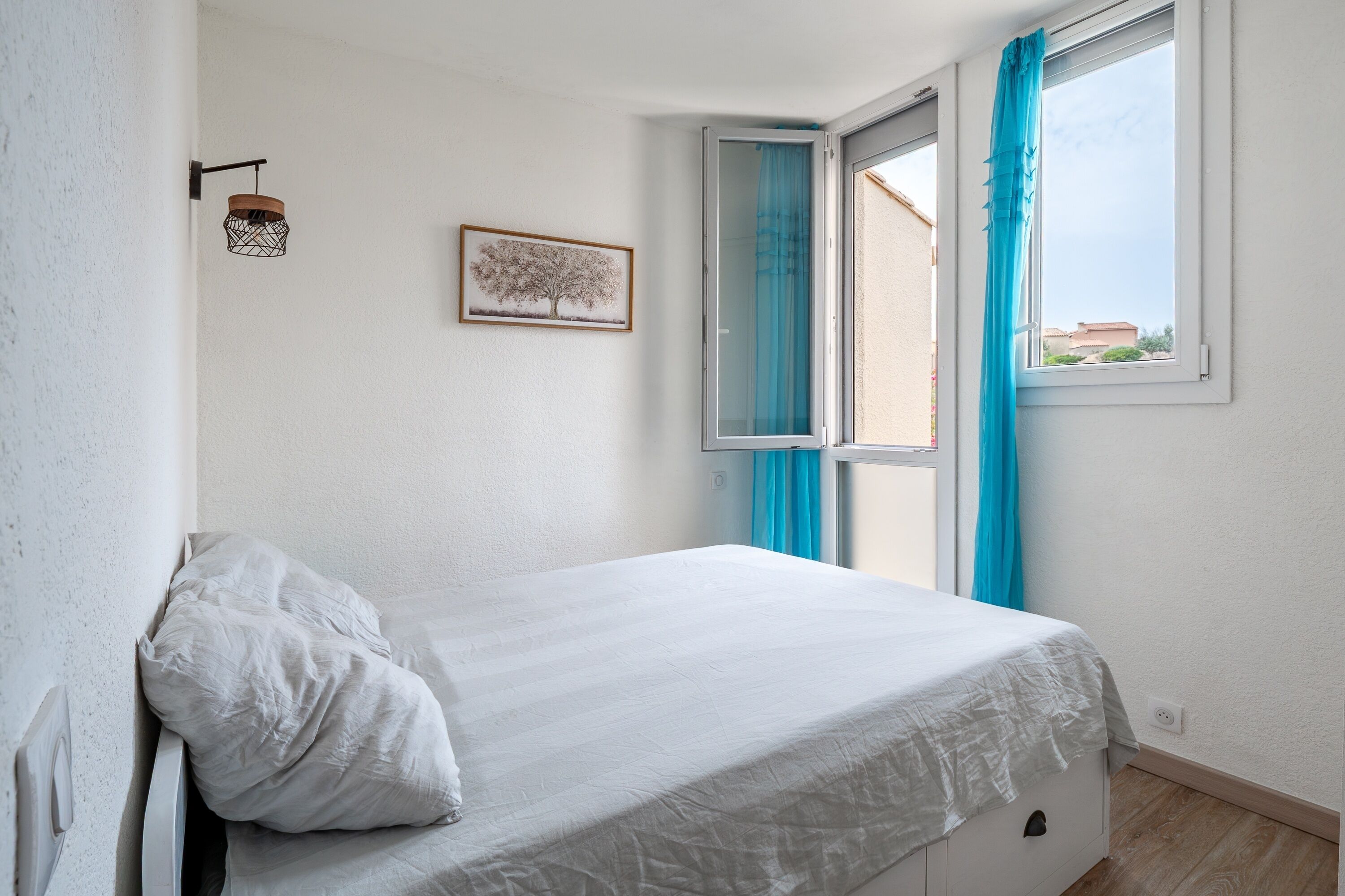 Un dormitorio con cama, ventana con cortinas, lámpara de pared e imagen enmarco.