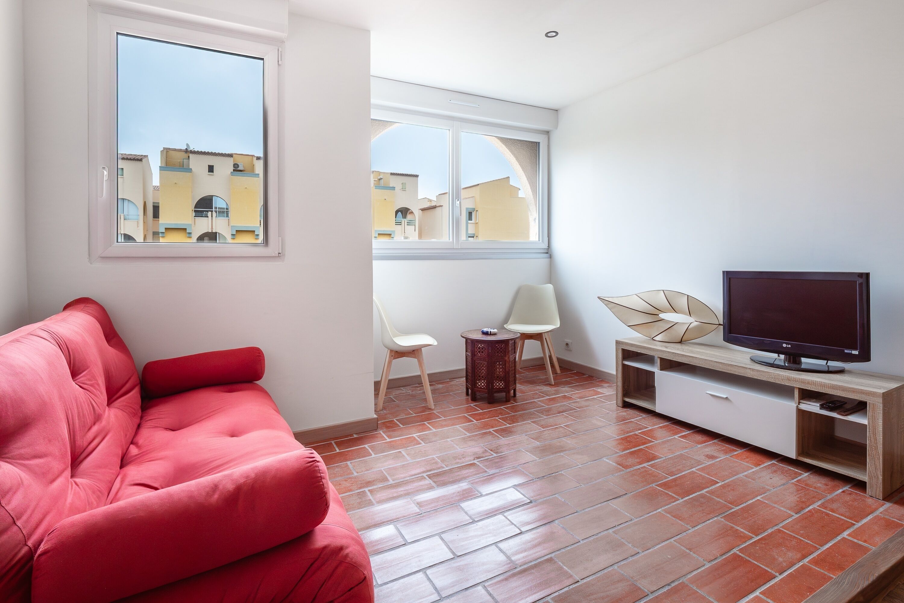 A modern living room with a red sofa, a TV on a wooden stand, and a view of buildings through large windows.