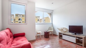 A modern living room with a red sofa, a TV on a wooden stand, and a view of buildings through large windows. - Le Maryland - Climatisé à 100 m de la plage (Leucate)