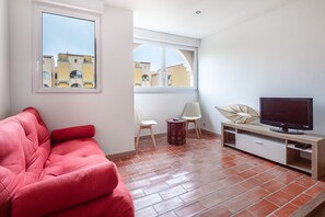 A modern living room with a red sofa, a TV on a wooden stand, and a view of buildings through large windows. - Le Maryland - Climatisé à 100 m de la plage (Leucate)