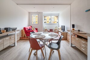A modern kitchen with a dining area, featuring a round table with placemats, wooden cabinets, and a red chair. - Le Maryland - Climatisé à 100 m de la plage (Leucate)