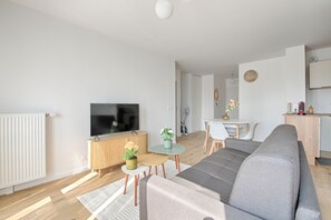 Una sala moderna con un televisor de pantalla plana grande, un sofá gris, una zona de comedor con una mesa y sillas pequeñas, y una cocina con microondas y cafetera.