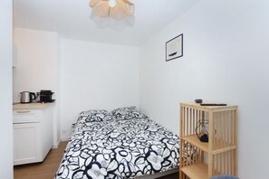 A small, neatly organized room with a bed, a wooden side table, a wall-mounted shelf, and a hanging light fixture. - Studio Océan - L’élégance d’une escale bretonne (Dol-de-Bretagne)