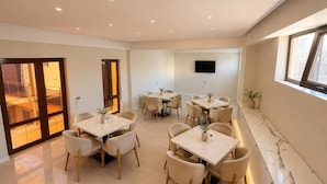 A modern dining area with multiple tables and chairs, a flat-screen TV mounted on the wall, and a large window providing natural light. - Darvaza Boutique Hotel (Bukhara)