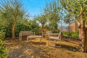An outdoor seating area with a bench and chairs, surrounded by trees and a fence.