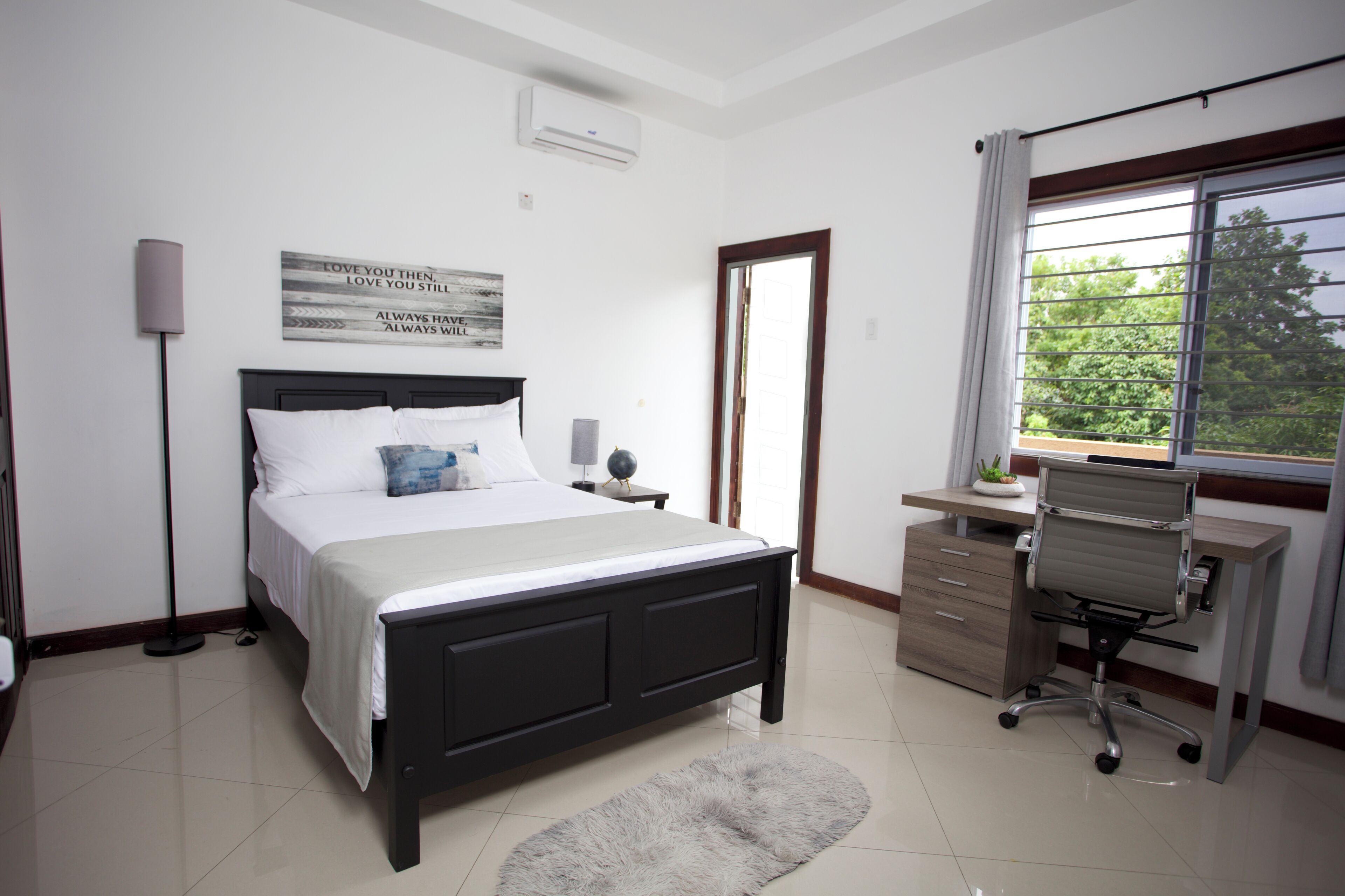 A bedroom with a bed, bedside lamp, desk, chair, and a window with a view.