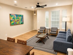 Een woonkamer met een houten eettafel, twee grijze fauteuils, een blauwe bank, een kleurrijk abstract schilderij en een plafondventilator.