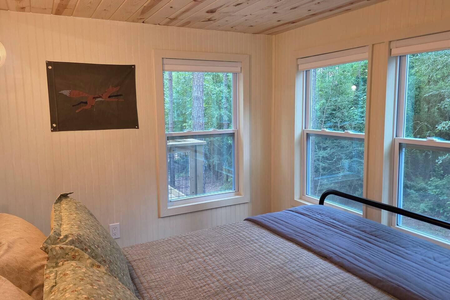 Un dormitorio con cama, almohada, un cuadro enmarcado en la pared y ventanas grandes con vista a árboles.