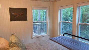 Un dormitorio con cama, almohada, un cuadro enmarcado en la pared y ventanas grandes con vista a árboles.