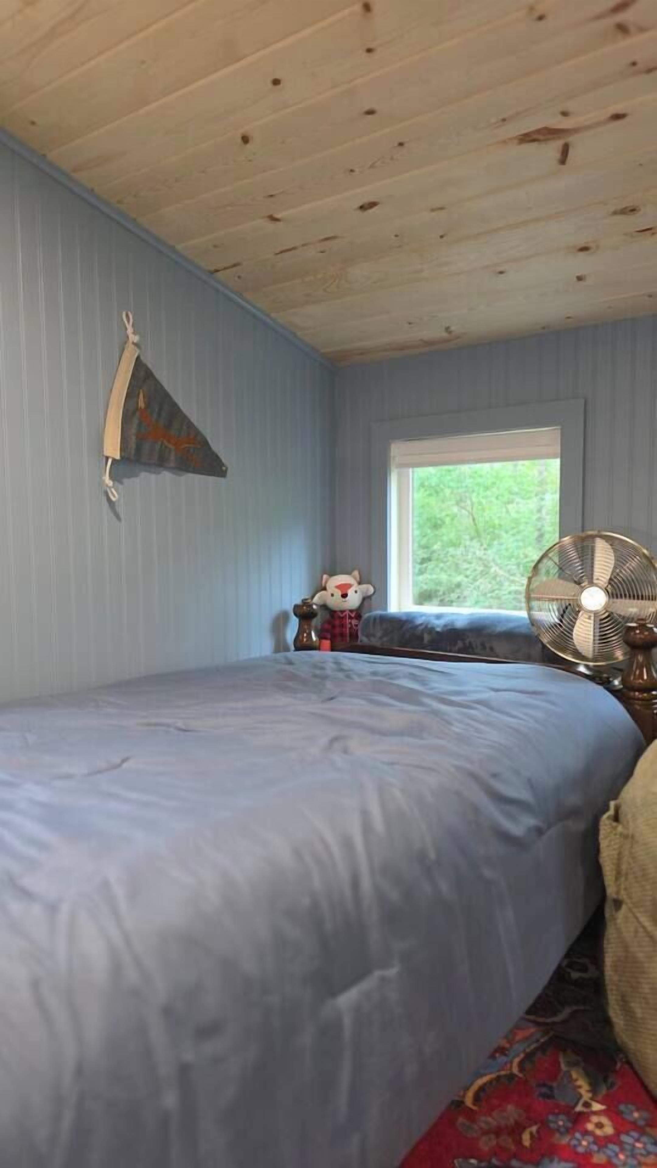 Un dormitorio con cama, ventana, ventilador y una mesita con un objeto decorativo.