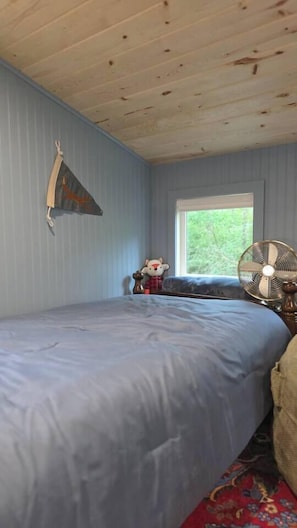 A bedroom with a bed, a window, a fan, and a small table with a decorative item.