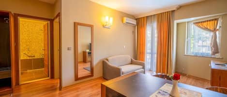 Habitación de hotel con piso de madera, un sofá, una mesa de comedor con un menú, un espejo, un equipo de aire acondicionado y una ventana con cortinas.