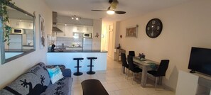 A modern living room with a sofa, a dining table with chairs, a kitchen area with cabinets and appliances, and a large wall clock. - Vacation Apartment 'Vacanze Mareverde' with Mountain View, Shared Pool and Wi-Fi (Costa Adeje)