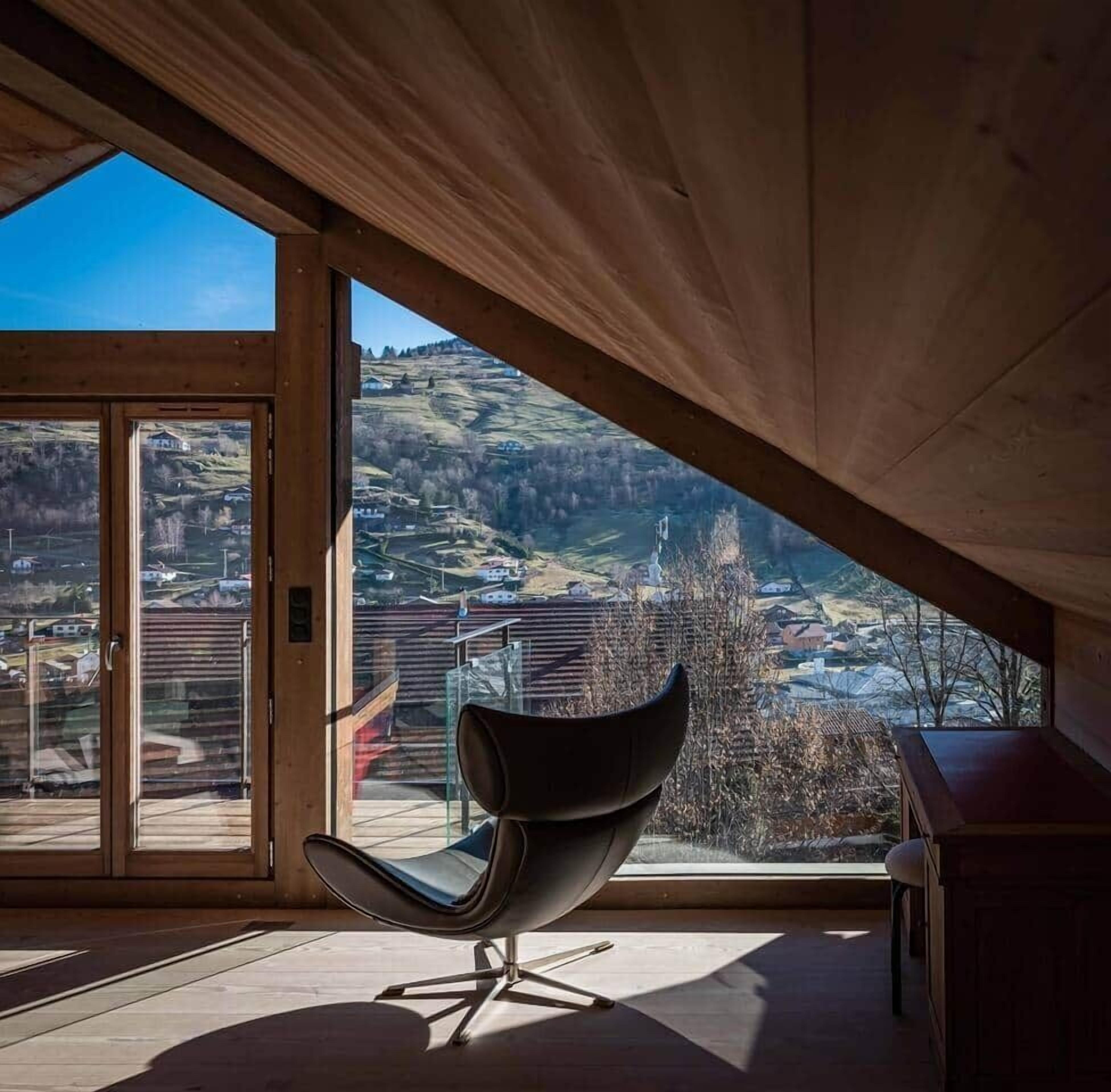 Ein modernes Interieur mit weitreichendem Panoramablick durch große Fenster, einem schwarzen Sessel und einer Holzbalkendecke.
