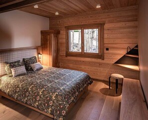 Een slaapkamer met een houten plafond, een groot bed met bloemenbeddelsels, een raam met uitzicht op bomen en een bankje.
