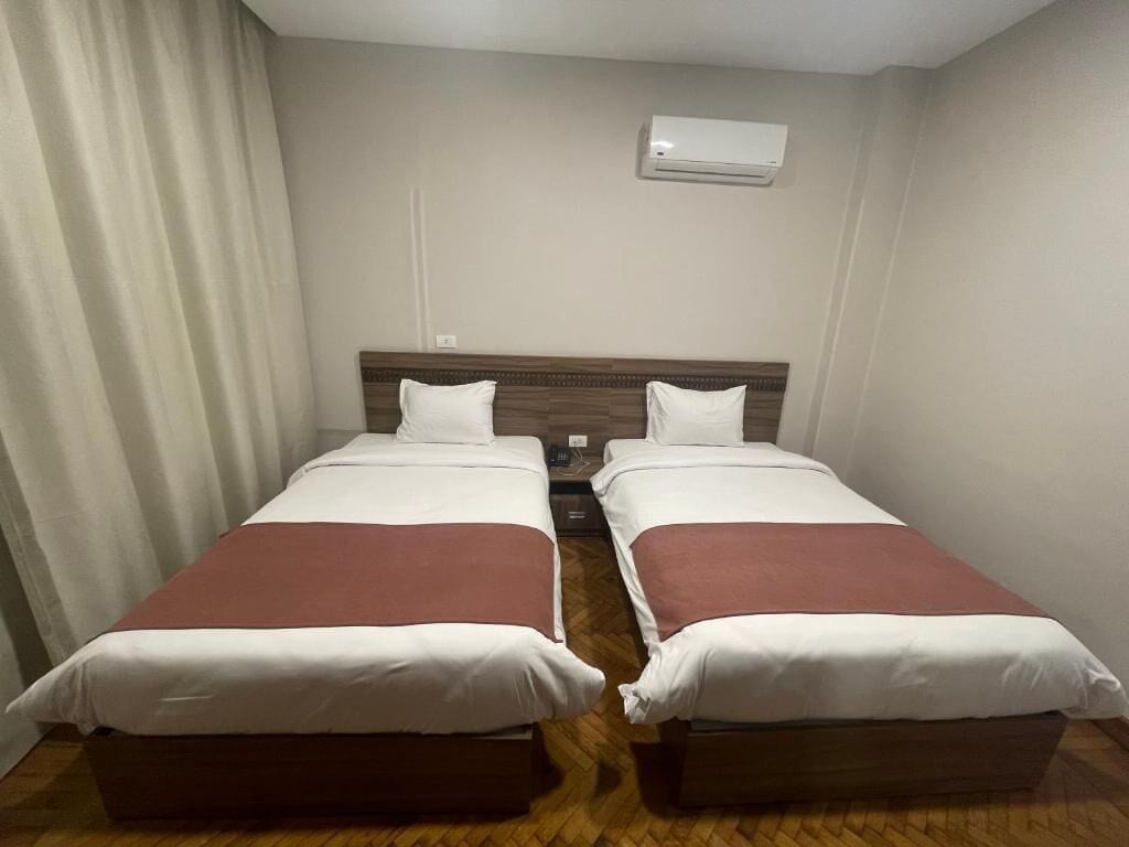 Une chambre avec deux lits, un plancher en bois, un climatiseur blanc fixé au mur et un rideau sur le côté gauche.