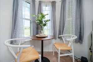 Une petite pièce avec une table ronde, deux chaises blanches, une plante en pot et des rideaux gris.