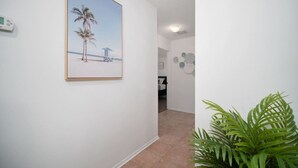 En hall med en inramad bild av en strand, en krukväxt och en dörr som leder till ett annat rum.