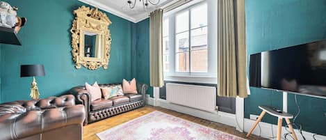 A room with teal walls, a large mirror, a leather sofa, a patterned rug, and a television.