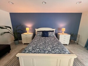 A bedroom with a bed, two bedside tables, a nightstand, a potted plant, and a wall-mounted shelf.