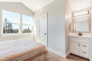 A bedroom with a bed, a window, a door, and a bathroom with a sink and mirror.