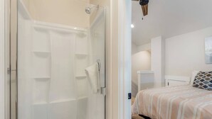 Een moderne slaapkamer met een douchecabine van glas, een bed met een gestreept beddegoed en een houten plafondventilator.