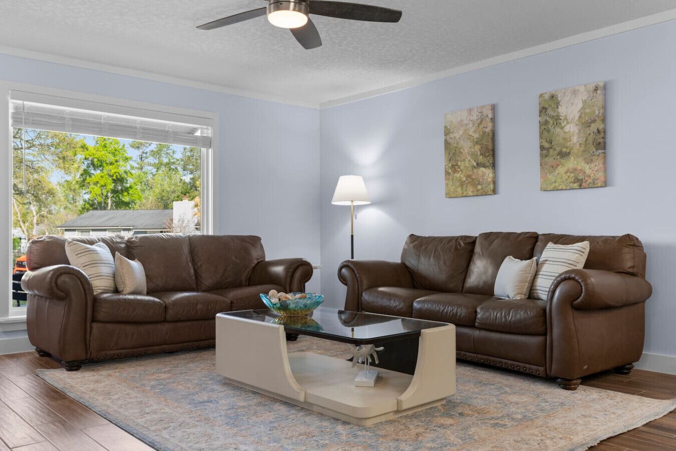 A living room with two brown leather sofas, a glass coffee table, a floor lamp, and abstract paintings on the wall.
