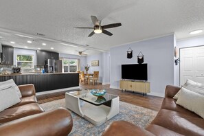A modern living room with a glass coffee table, leather sofas, a TV, and a kitchen in the background. - 10 Min to Mayo & Beach • Sleeps 5 • Clean & Quiet (Jacksonville)