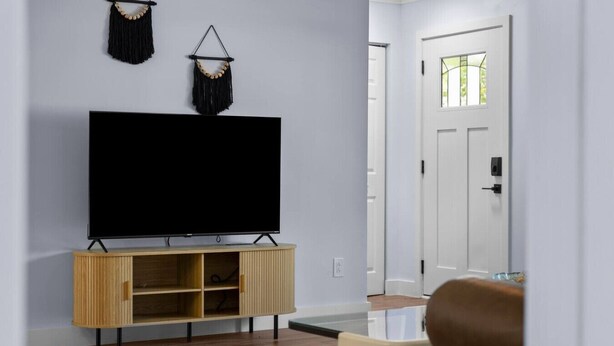 A modern living room with a flat-screen TV on a wooden stand, a white door in the background, and a brown sofa. - San Pablo Retreat Near Mayo Clinic Mins to Beaches (Jacksonville)