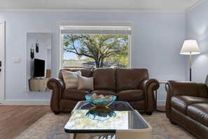Een woonkamer met een bruine leren bank, een glazen salontafel met een decoratieve kom, een televisie op een houten standaard, en een groot raam met uitzicht op bomen.