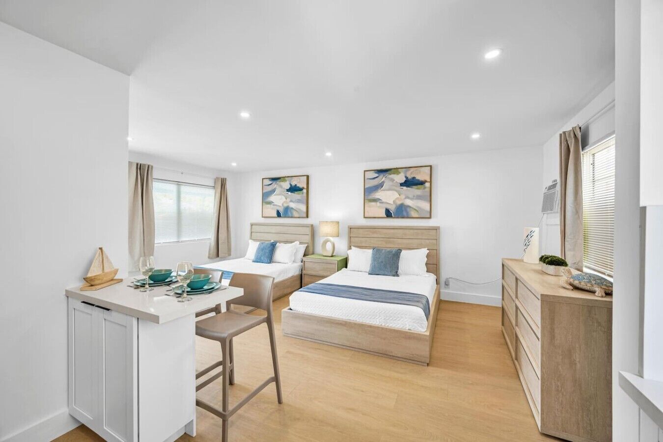A modern hotel room with two beds, a small kitchenette, and a dining area.