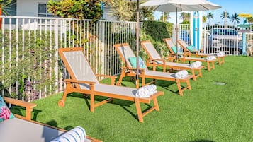 Une rangée de fauteuils de jardin en bois, dotés de coussins, est disposée sur une pelouse verdoyante, avec une clôture blanche et des plantes tropicales en arrière-plan.